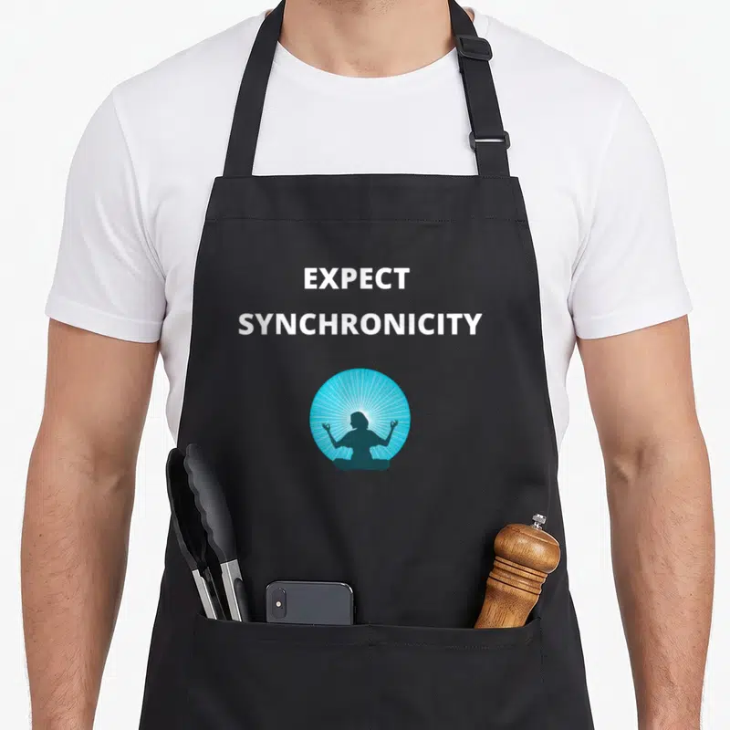 Expect manifestation apron - White expect synchronicity text above teal lotus figure in blue sunburst circle — shown on a black cotton canvas apron against a white background