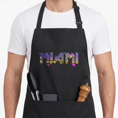 Skyline florida pride apron - MIAMI text formed by negative space containing purple pink yellow and grey city buildings — shown on a black cotton canvas apron against a white background