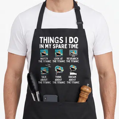 Rms birthday apron - Cartoon Titanic ship scenes with text Watch Look Research Talk Think Dream About The Titanic — shown on a black cotton canvas apron against a white background