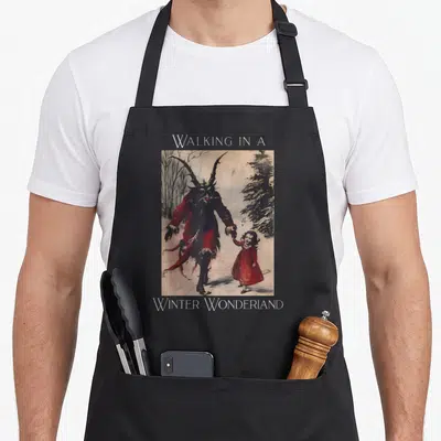 Walking winter apron - Horned monster holding girl hand in snow with Walking In A Winter Wonderland text — shown on a black cotton canvas apron against a white background