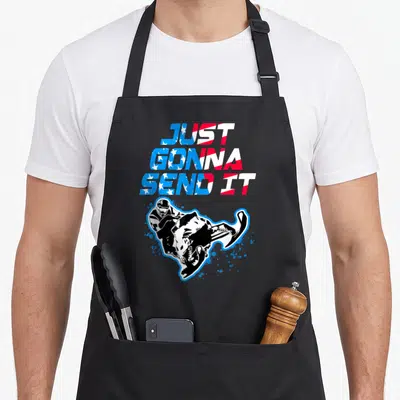 Flag snowmobile girl apron - Stacked flag text JUST GONNA SEND IT above cartoon snowmobiler with blue splatters — shown on a black cotton canvas apron against a white background