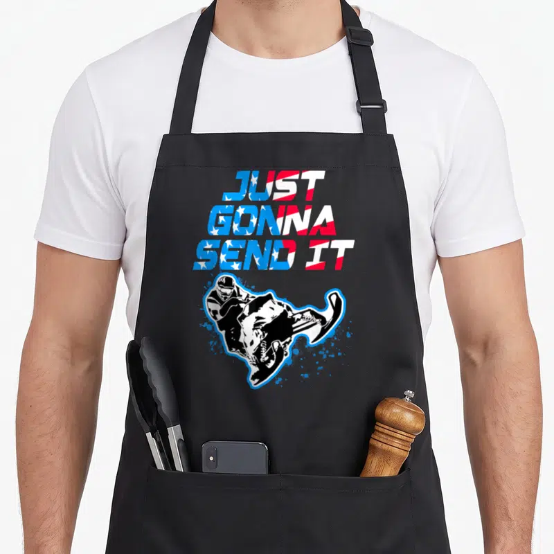 Flag snowmobile girl apron - Stacked flag text JUST GONNA SEND IT above cartoon snowmobiler with blue splatters — shown on a black cotton canvas apron against a white background