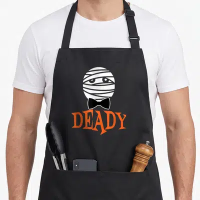 Deady mummy and me aprons - Cartoon mummy head with black bow tie above orange DEADY text — shown on a black cotton canvas apron against a white background