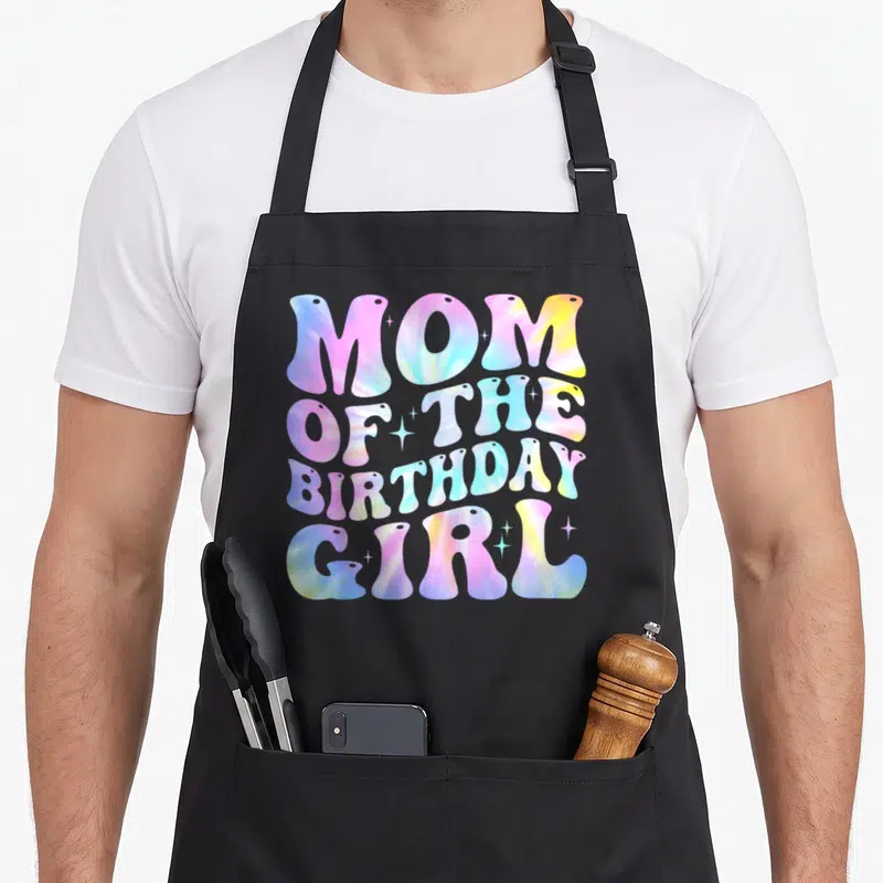 Colourful tie apron - Tie-dye text reading Mom of the Birthday Girl in pink blue yellow and purple with white sparkles — shown on a black cotton canvas apron against a white background