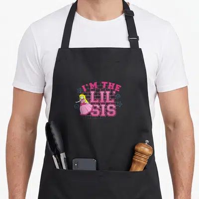 Lil princess apron - Princess Peach character beside pink I'M THE LIL' SIS text with blue stars and brick background — shown on a black cotton canvas apron against a white background