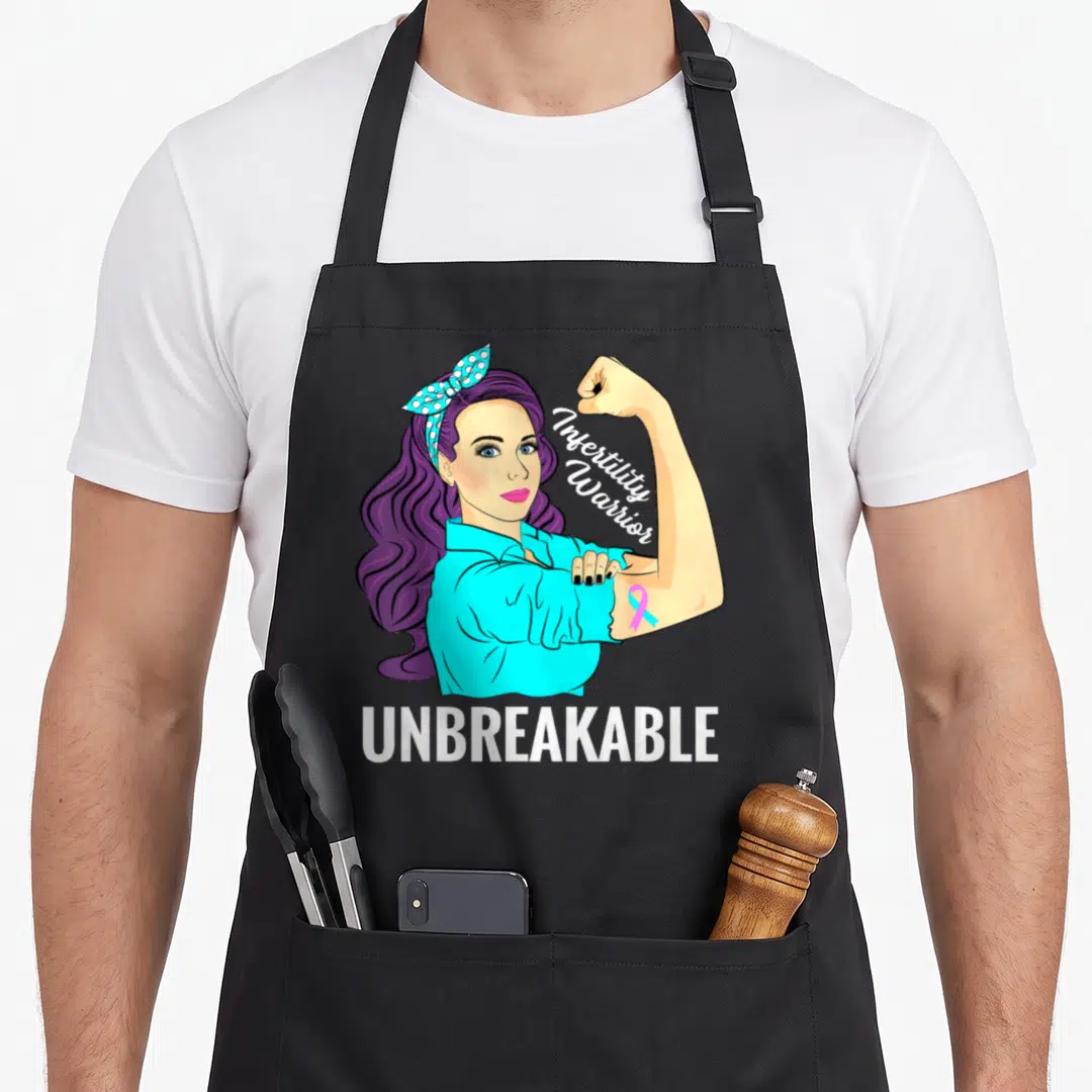 Warrior infertility awareness apron - Cartoon woman flexing arm with Infertility Warrior text and pink cyan ribbon — shown on a black cotton canvas apron against a white background
