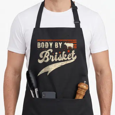 Funny brisket body apron - Distressed cow silhouette and BODY BY Brisket text with orange lines and stars — shown on a black cotton canvas apron against a white background