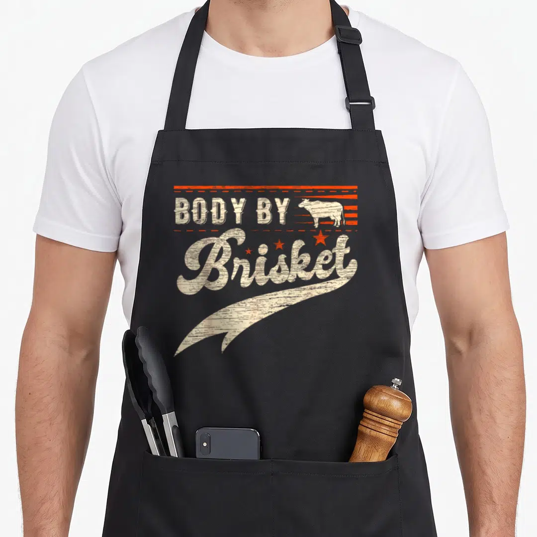 Funny brisket body apron - Distressed cow silhouette and BODY BY Brisket text with orange lines and stars — shown on a black cotton canvas apron against a white background