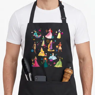 Magical princess apron - Scattered cartoon Disney princesses with icons in pink yellow blue green purple — shown on a black cotton canvas apron against a white background