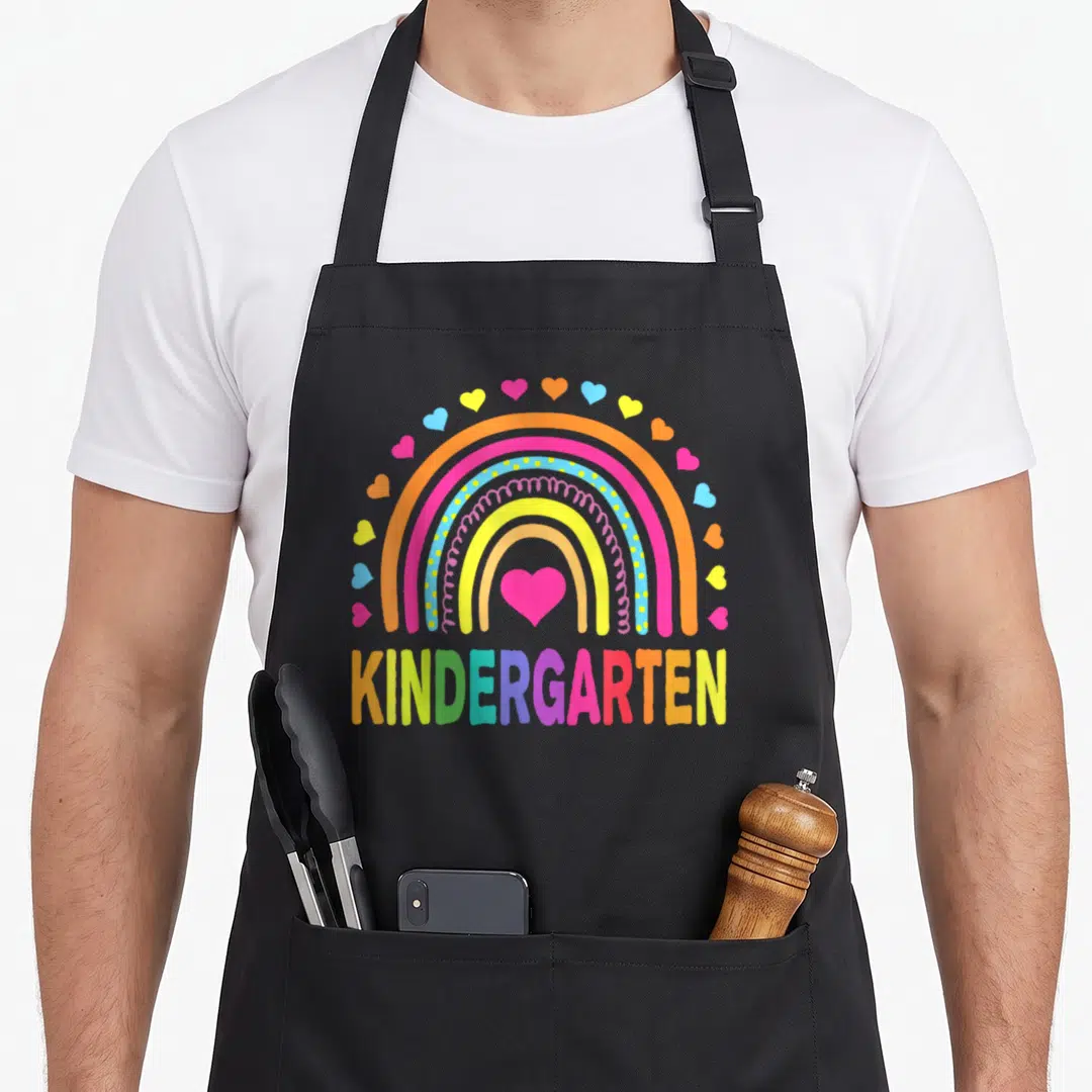 Rainbow cute teacher aprons - Multi-colored rainbow with hearts above the word KINDERGARTEN in cartoon lettering — shown on a black cotton canvas apron against a white background