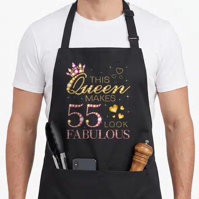 Fabulous 55th birthday apron - Gold and pink rhinestone style text reading THIS Queen MAKES 55 LOOK FABULOUS with crown — shown on a black cotton canvas apron against a white background