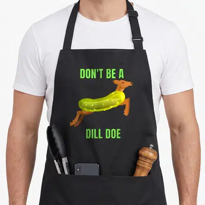 Dill doe pickle apron - Cartoon pickle deer with green text reading DON'T BE A DILL DOE — shown on a black cotton canvas apron against a white background