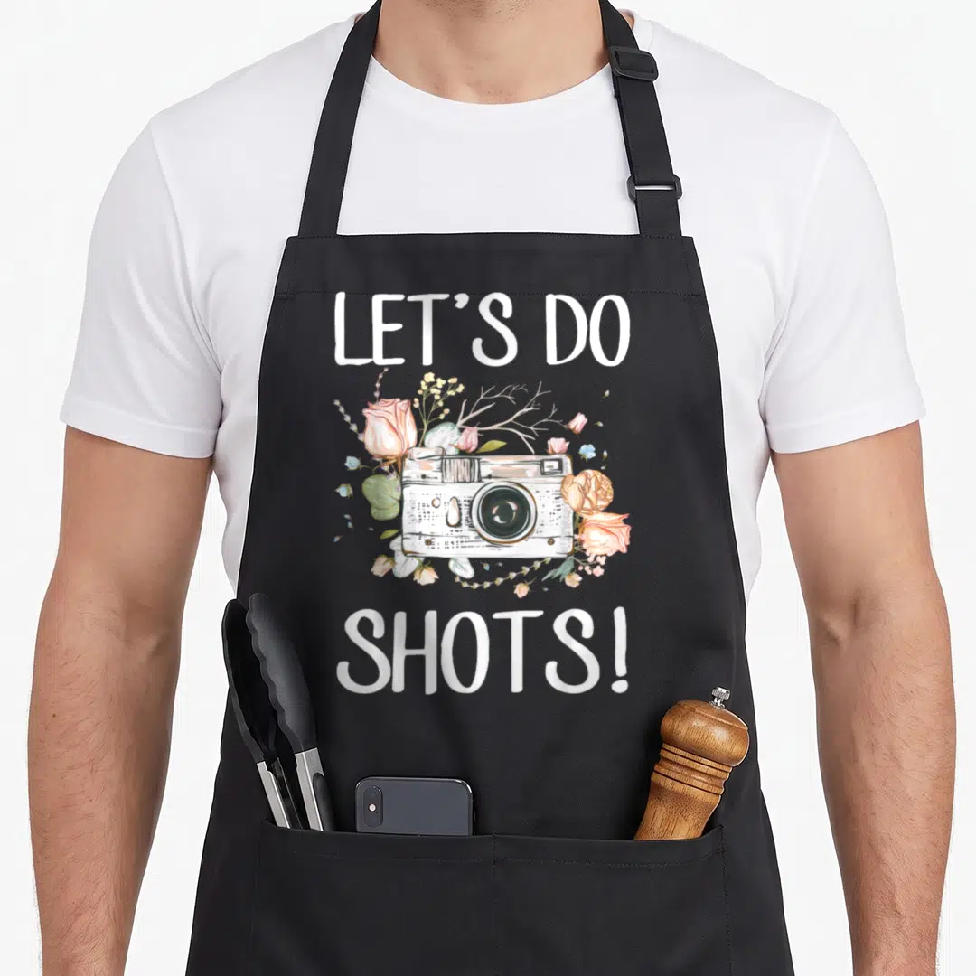 Dress photographer apron - Watercolor camera with pink roses and LET'S DO SHOTS text — shown on a black cotton canvas apron against a white background