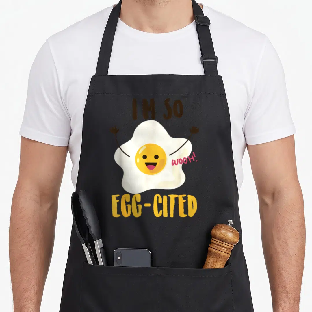 Cited pun egg apron - Cartoon fried egg with raised arms and text I'M SO WOOH EGG-CITED in white yellow brown and pink — shown on a black cotton canvas apron against a white background