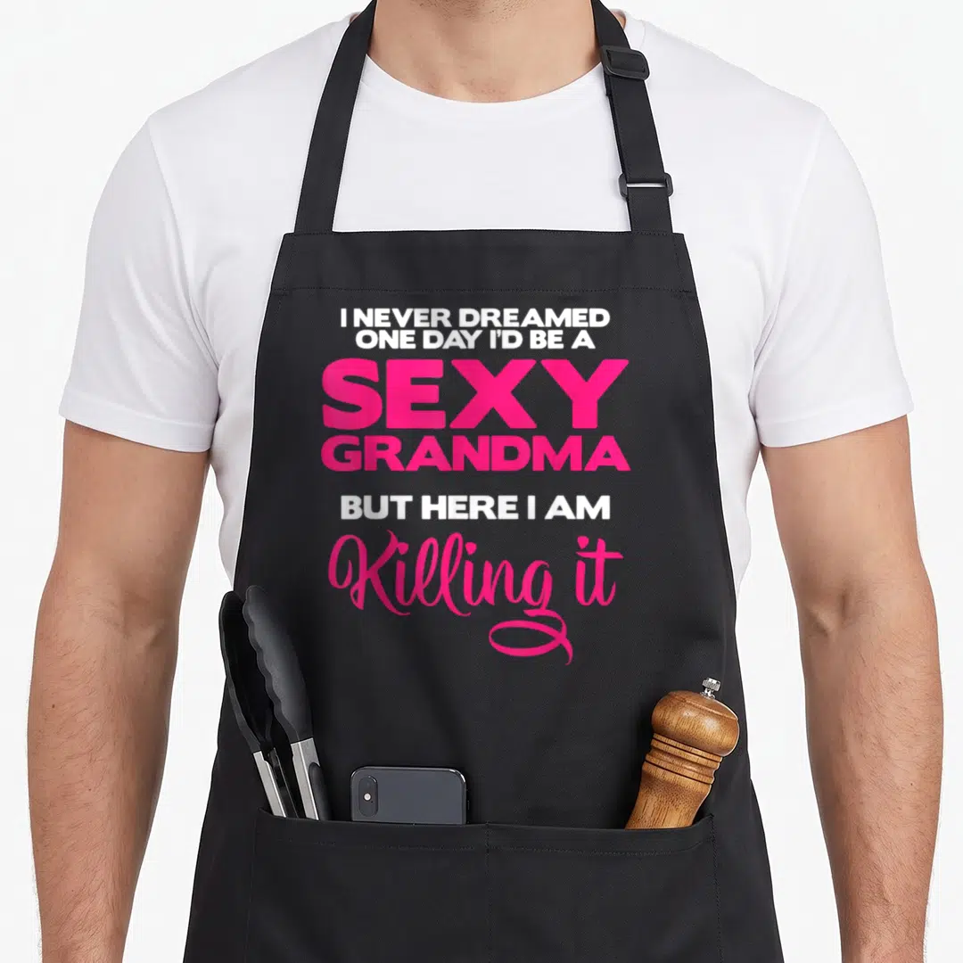 Grandma chef outfit apron - Stacked white and pink typography reading I never dreamed one day I'd be a sexy grandma — shown on a black cotton canvas apron against a white background
