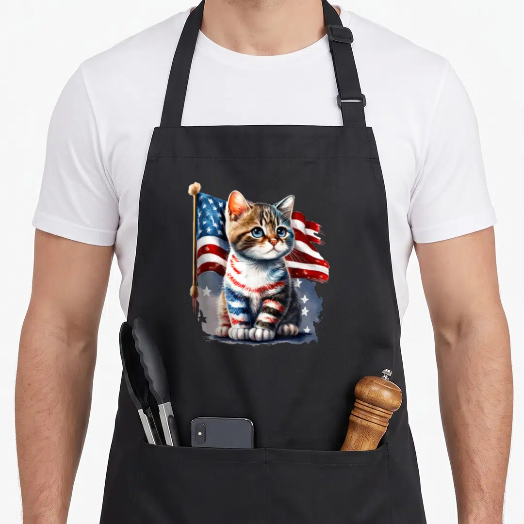 Memorial cat 4th of july apron - Realistic tabby kitten with red white blue striped fur sitting in front of waving US — shown on a black cotton canvas apron against a white background