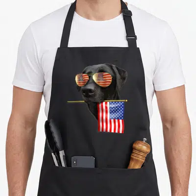 Labrador 4th of july apron - Realistic black lab holding a flag with sunglasses reflecting stars and stripes — shown on a black cotton canvas apron against a white background