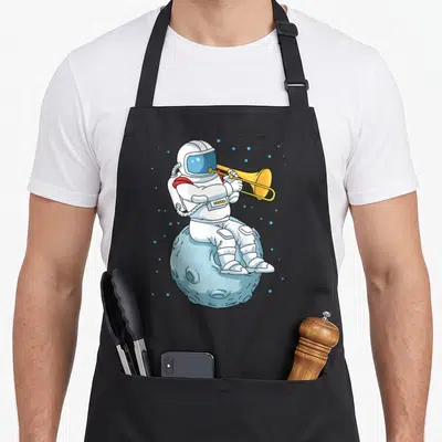 Trombone neat apron - Cartoon astronaut in white suit playing yellow trumpet while sitting on light blue moon — shown on a black cotton canvas apron against a white background