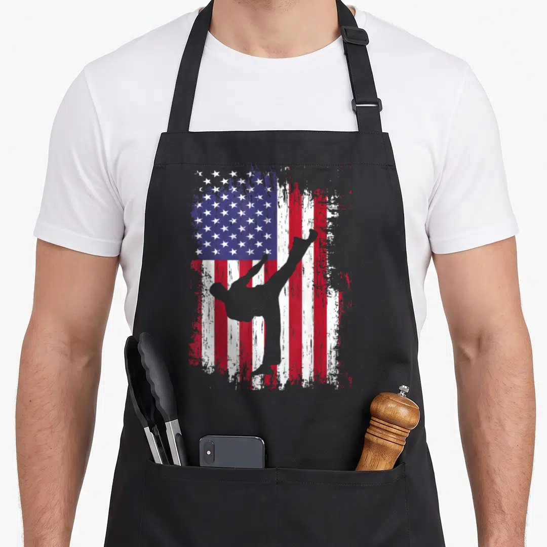 Funny taekwondo karate apron - Grey martial artist silhouette kicking in front of a distressed red white and blue USA — shown on a black cotton canvas apron against a white background