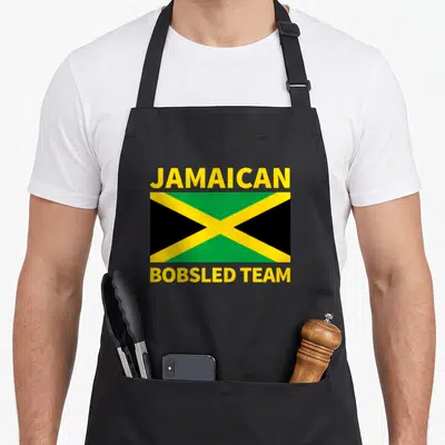 Jamaican halloween apron - JAMAICAN BOBSLED TEAM text surrounding yellow green and black flag — shown on a black cotton canvas apron against a white background