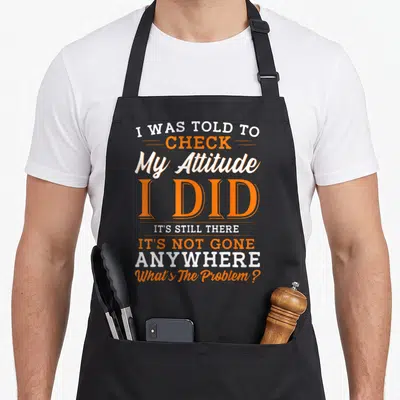 Sarcasm attitude aprons - Stacked orange and white typography reading I Was Told To Check My Attitude I Did It's Still — shown on a black cotton canvas apron against a white background