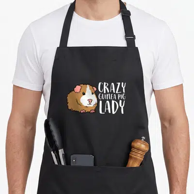 Vintage guinea pet apron - Cartoon guinea pig with white brown pink fur next to stacked text CRAZY GUINEA PIG LADY — shown on a black cotton canvas apron against a white background