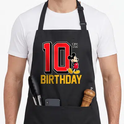 Funny 10th birthday apron - Mickey Mouse next to large red 10TH BIRTHDAY text in yellow block — shown on a black cotton canvas apron against a white background