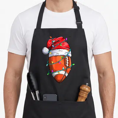 Football christmas santa apron - Cartoon football wearing a red Santa hat wrapped in multicolored Christmas lights — shown on a black cotton canvas apron against a white background