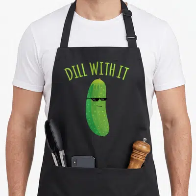Dill pickle apron - Green cartoon pickle wearing black pixelated sunglasses under arched DILL WITH IT text — shown on a black cotton canvas apron against a white background