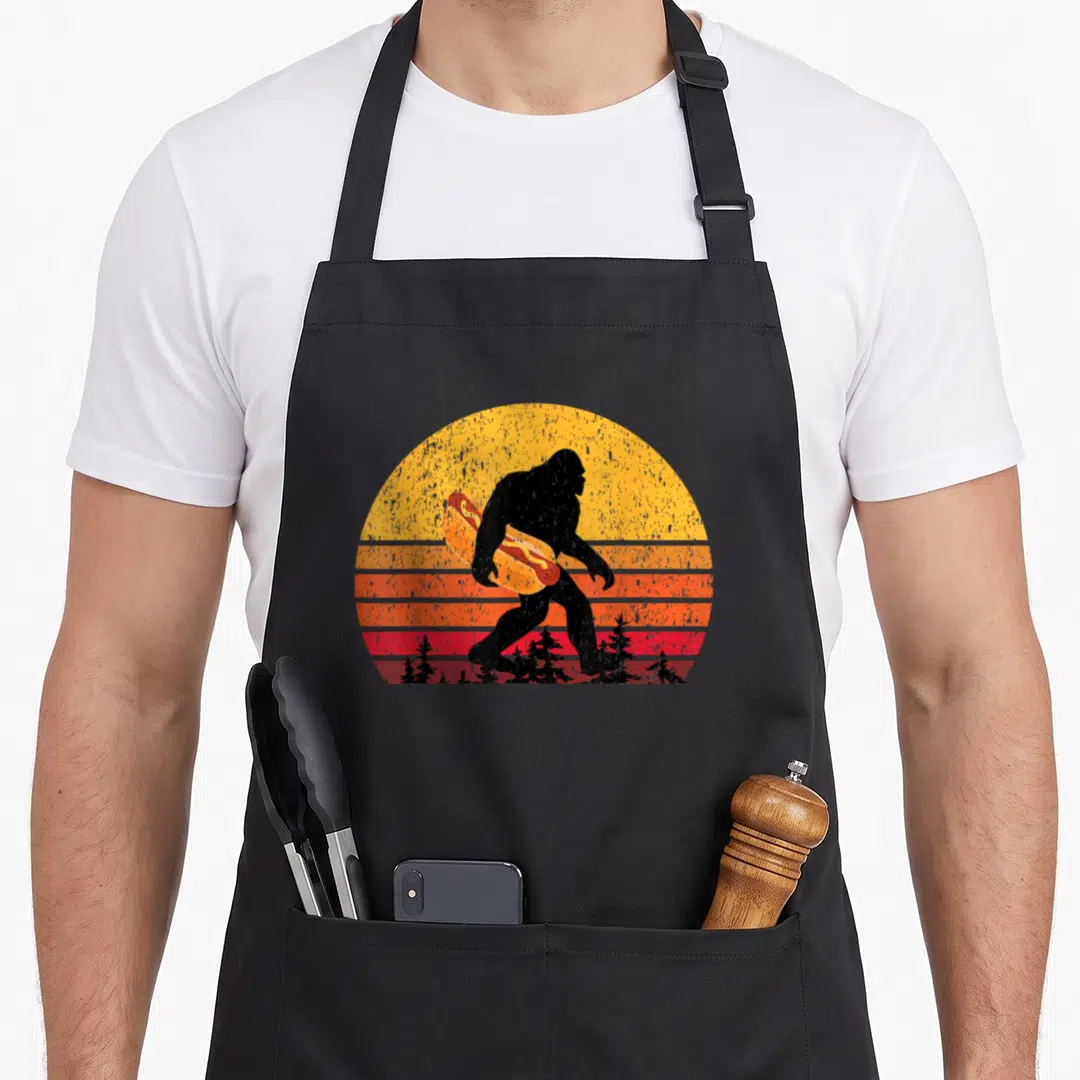 Bigfoot hot dog apron - Black Bigfoot silhouette holding giant hot dog against yellow orange red striped sun — shown on a black cotton canvas apron against a white background