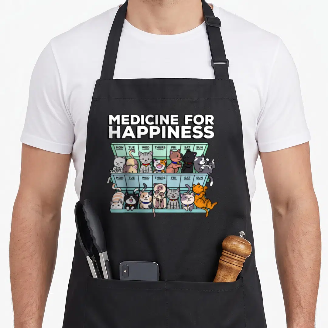 Medicine kitten apron - Cartoon cats in a weekly pill organizer labeled Monday through Sunday mornings and evenings — shown on a black cotton canvas apron against a white background