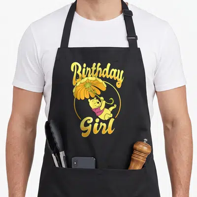 Funny birthday floral aprons - Winnie the Pooh holding a yellow flower umbrella with Birthday Girl text in yellow script — shown on a black cotton canvas apron against a white background
