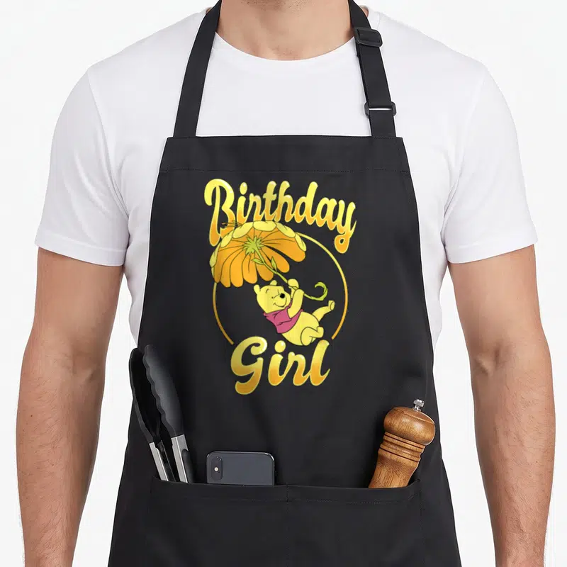Funny birthday floral aprons - Winnie the Pooh holding a yellow flower umbrella with Birthday Girl text in yellow script — shown on a black cotton canvas apron against a white background