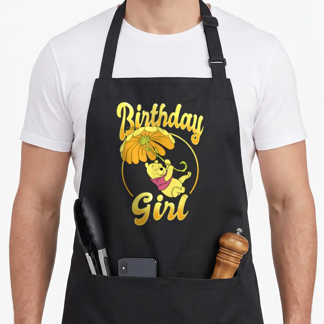 Funny birthday floral aprons - Winnie the Pooh holding a yellow flower umbrella with Birthday Girl text in yellow script — shown on a black cotton canvas apron against a white background