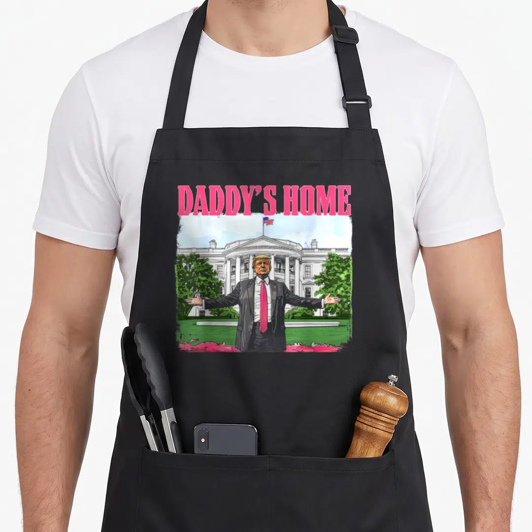 Trump daddy apron - DADDY'S HOME text above cartoon Trump in front of White House with pink and green accents — shown on a black cotton canvas apron against a white background