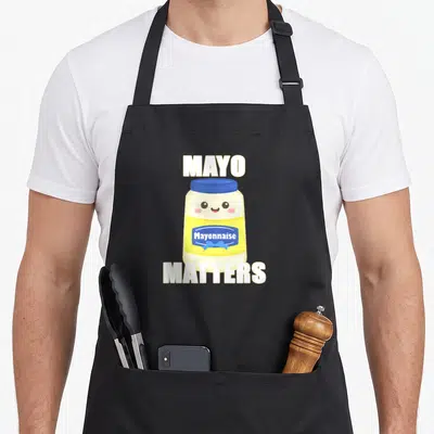 Mayo funny jam apron - Smiling cartoon mayonnaise jar with blue lid, yellow label, and white MAYO MATTERS text — shown on a black cotton canvas apron against a white background