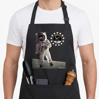July moon apollo apron - Astronaut on moon surface next to Apollo 11 mission patch with circle of stars — shown on a black cotton canvas apron against a white background