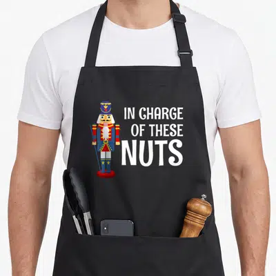 Costume nutcracker apron - Cartoon nutcracker soldier next to white text reading IN CHARGE OF THESE NUTS — shown on a black cotton canvas apron against a white background