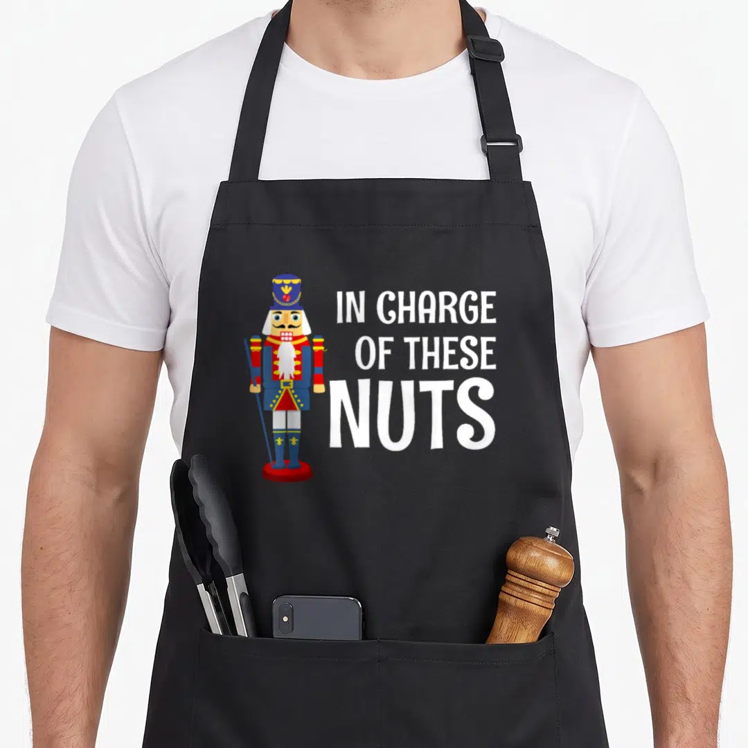 Costume nutcracker apron - Cartoon nutcracker soldier next to white text reading IN CHARGE OF THESE NUTS — shown on a black cotton canvas apron against a white background