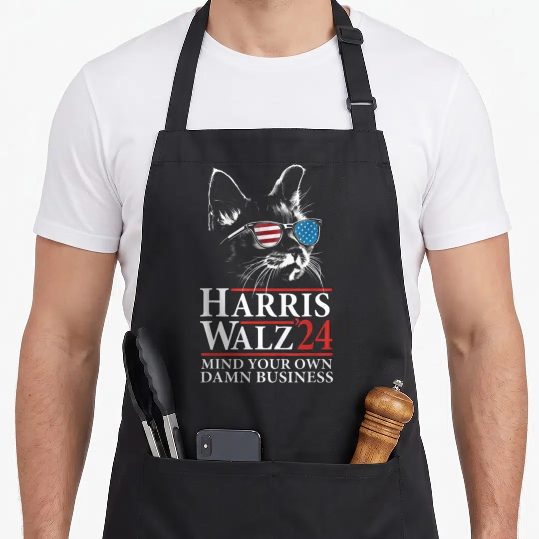 Walz cat lady apron - Cat wearing flag sunglasses with Harris Walz 24 and Mind Your Own Damn Business text — shown on a black cotton canvas apron against a white background