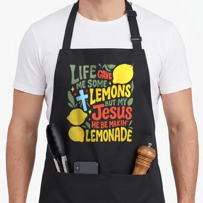 Jesus lemonade apron - LIFE GAVE ME SOME LEMONS text with cartoon cross and yellow lemons on black background — shown on a black cotton canvas apron against a white background