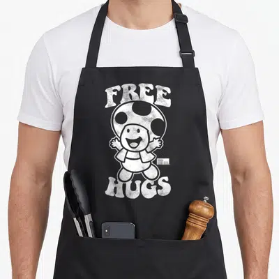 Free toad apron - White cartoon Toad character with Free Hugs text and small Super Mario Bros logo — shown on a black cotton canvas apron against a white background