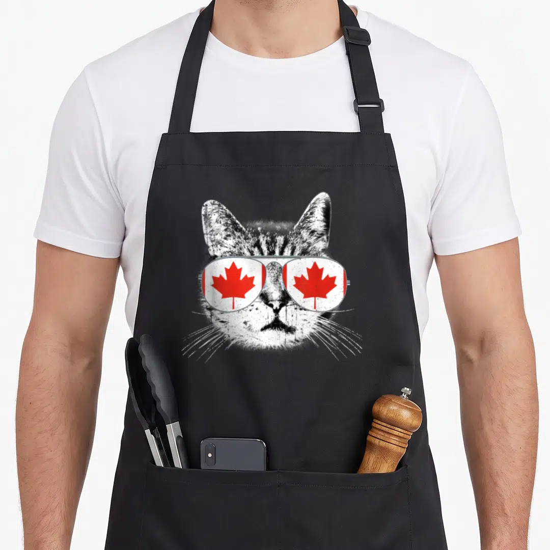 Canada cat apron - Distressed black and white cat head wearing aviator sunglasses with red maple leaf lenses — shown on a black cotton canvas apron against a white background