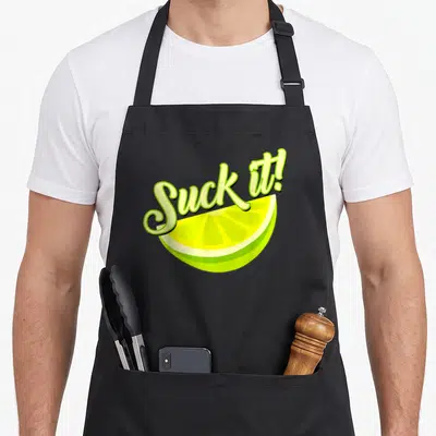 Suck lemon apron - Cartoon lime slice with Suck it script text in green and yellow — shown on a black cotton canvas apron against a white background