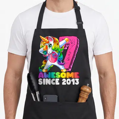 Unicorn 7th birthday apron - Dabbing unicorn with rainbow mane next to pink number 7 and AWESOME SINCE 2013 text — shown on a black cotton canvas apron against a white background