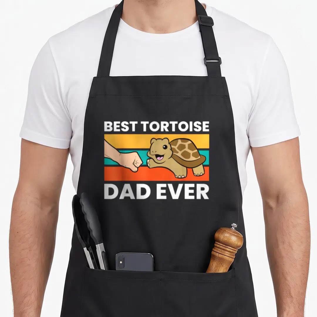 Funny tortoise turtle dad apron - Cartoon tortoise fist-bumping human hand over yellow teal orange and red stripes — shown on a black cotton canvas apron against a white background