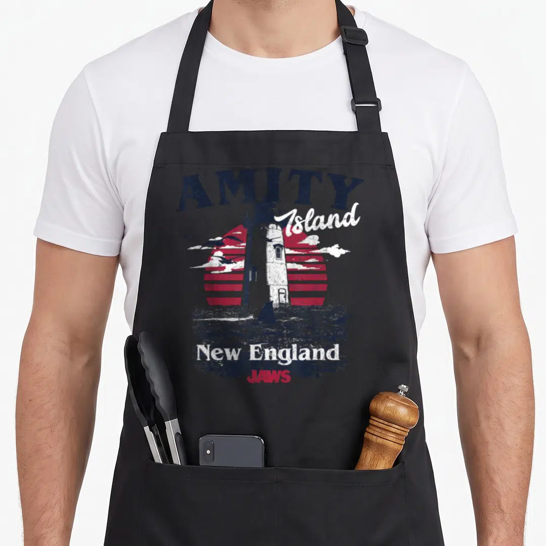 Amity england apron - Retro lighthouse with JAWS text and shark fin on red striped sun — shown on a black cotton canvas apron against a white background