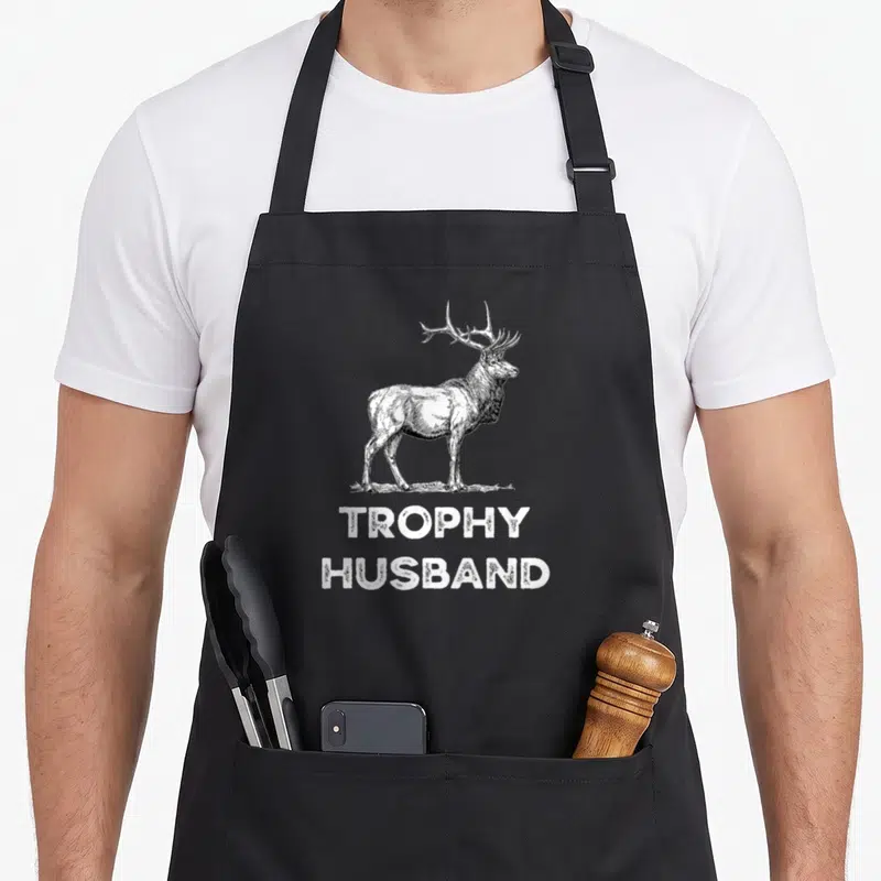 Trophy husband father day apron - Realistic white deer illustration above stacked TROPHY HUSBAND text on grey background — shown on a black cotton canvas apron against a white background