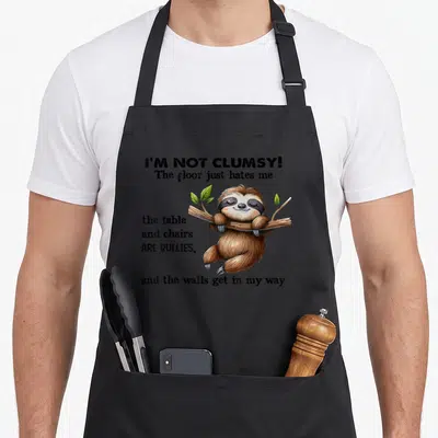 Sloth table apron - Cartoon sloth hanging from branch with text about floor hatred and table bullying — shown on a black cotton canvas apron against a white background