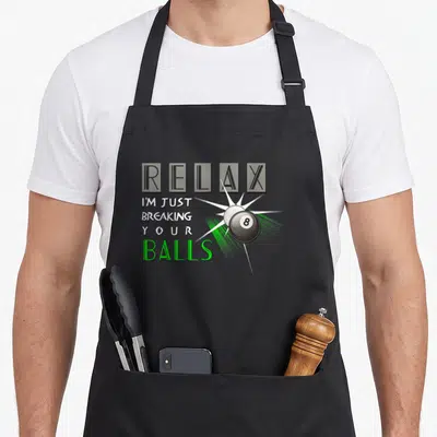 Breaking billiards lover apron - RELAX I'M JUST BREAKING YOUR BALLS text next to cartoon 8-ball collision in green — shown on a black cotton canvas apron against a white background